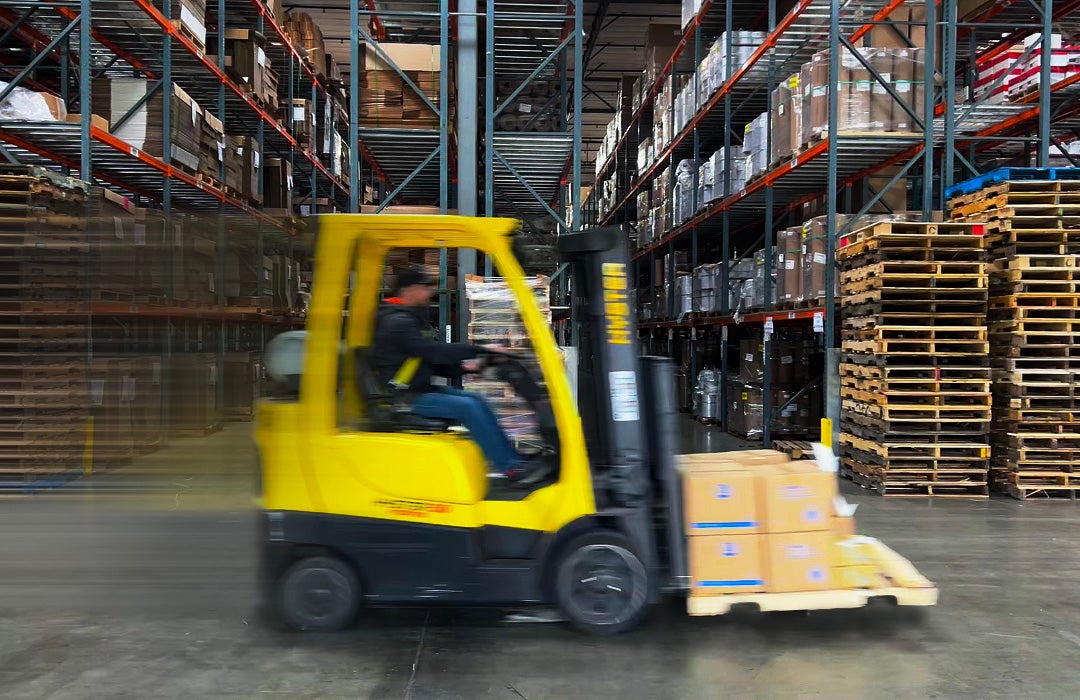 Yellow forklift driving through a warehouse in front of a pile of wooden pallets